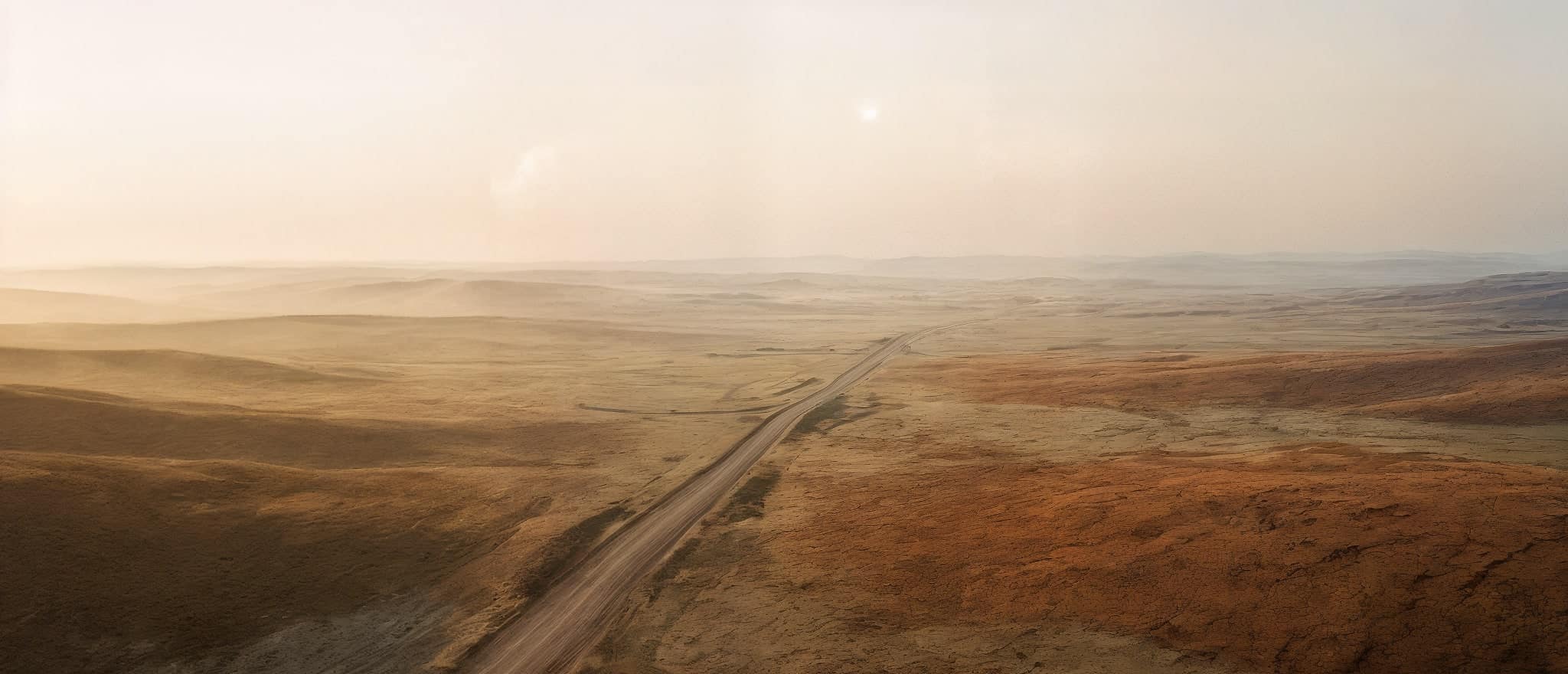Hazy desert plain with a single road running to the horizon under a soft sun